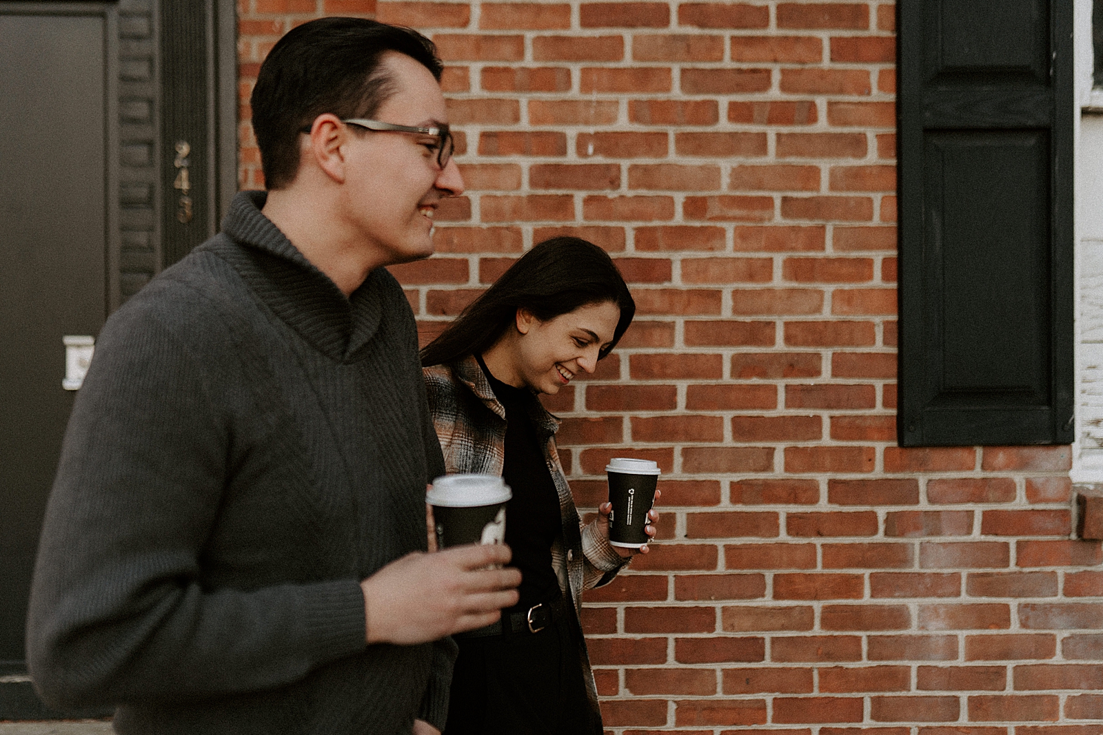 Couple looking at each other next to red brick building and holding coffees