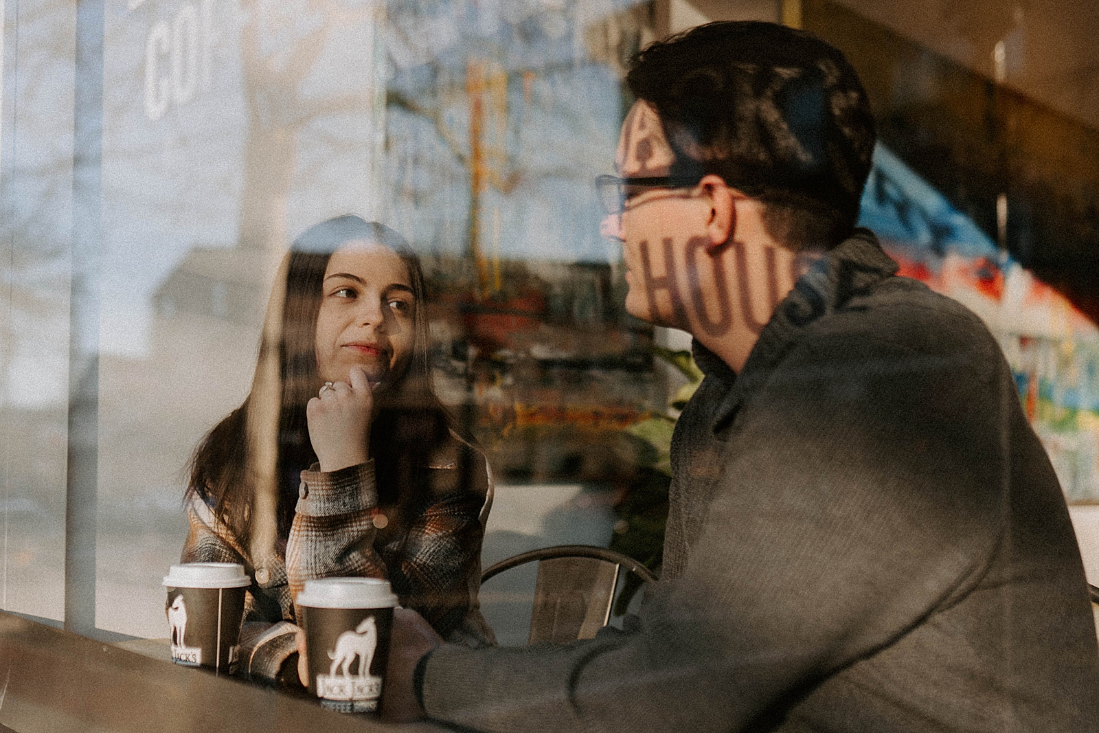 Through glass window portrait of couple sitting together and looking at each other with coffee